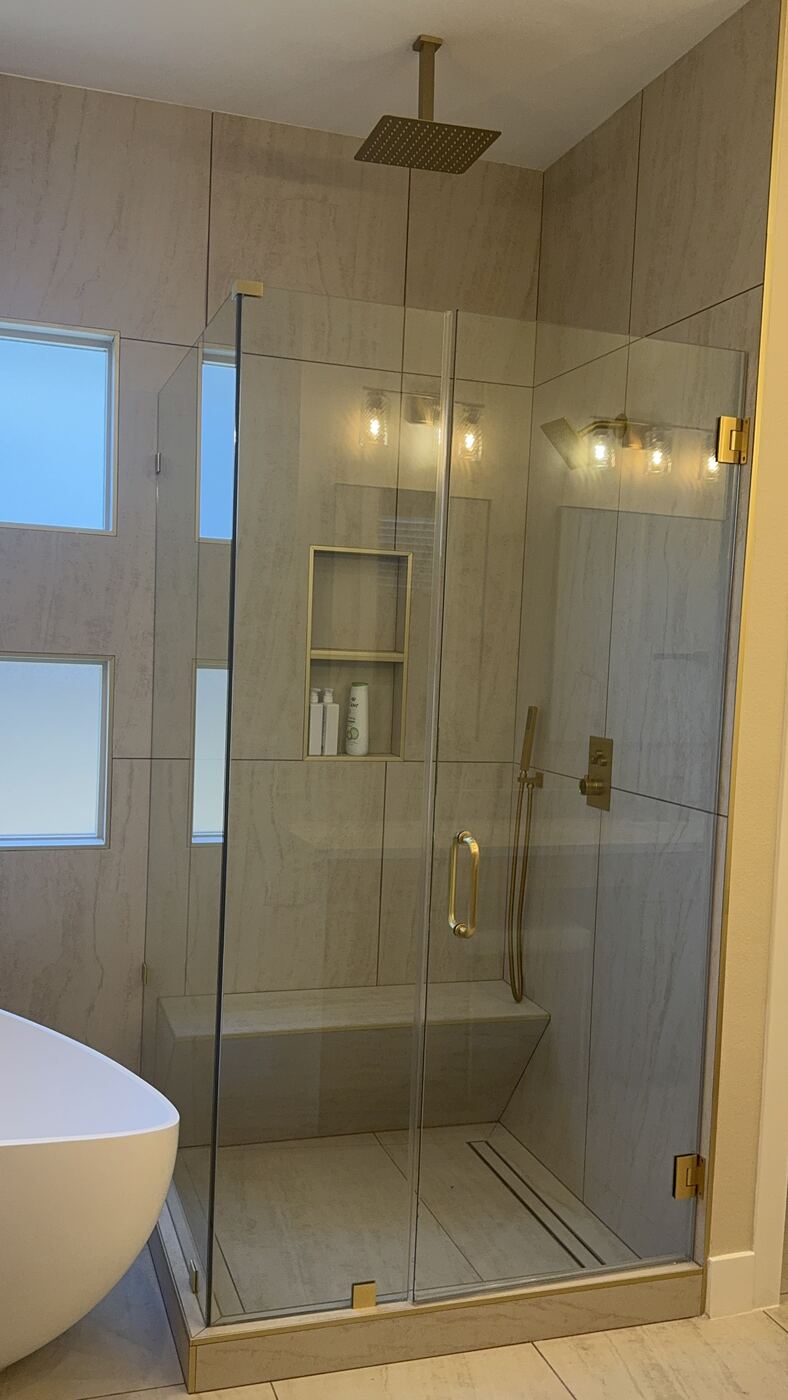 Frameless glass shower with brushed gold rain shower head and tiled niche in a primary bathroom remodel
